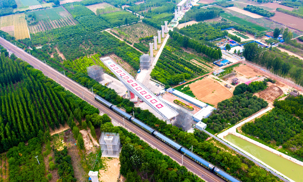 跨京九鐵路連續(xù)梁是雄安機(jī)場(chǎng)快線首座涉及運(yùn)營(yíng)鐵路的轉(zhuǎn)體梁，所跨京九鐵路每日運(yùn)行列車多達(dá)百余列。為確保轉(zhuǎn)體期間京九鐵路安全運(yùn)營(yíng)，中鐵四局多次組織專家評(píng)審、優(yōu)化施工方案，并在正式轉(zhuǎn)體前試轉(zhuǎn)，通過稱重平衡試驗(yàn)測(cè)試轉(zhuǎn)體部分的不平衡力矩、偏心矩、摩阻力矩及摩擦系數(shù)等參數(shù)，以實(shí)現(xiàn)橋梁轉(zhuǎn)體的配重要求，在轉(zhuǎn)體過程中，根據(jù)轉(zhuǎn)臺(tái)上標(biāo)識(shí)的轉(zhuǎn)角刻度及梁面軸線實(shí)行嚴(yán)格監(jiān)控。圖為跨京九鐵路轉(zhuǎn)體梁轉(zhuǎn)體前航拍。