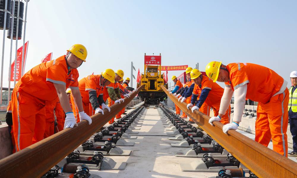 為優(yōu)質(zhì)高效完成鋪軌任務(wù)，建設(shè)者提前在位于荊州站北側(cè)的鋪軌基地儲存長鋼軌，以鋪軌基地為起點(diǎn)，采用TC-500型鋪軌機(jī)由荊州站一路向北鋪設(shè)長鋼軌；加持智能行車調(diào)度指揮系統(tǒng)，全天候?qū)︿佨墮C(jī)、長軌運(yùn)輸車、焊軌車等設(shè)備的運(yùn)行狀態(tài)及作業(yè)位置進(jìn)行監(jiān)控和統(tǒng)一調(diào)度指揮，保證了交叉施工安全有序，并且提高了鋪軌速度，每天單向鋪軌可達(dá)6公里。圖為鋪軌作業(yè)現(xiàn)場，作業(yè)人員在鋪軌機(jī)的牽引下正在安裝軌道。
