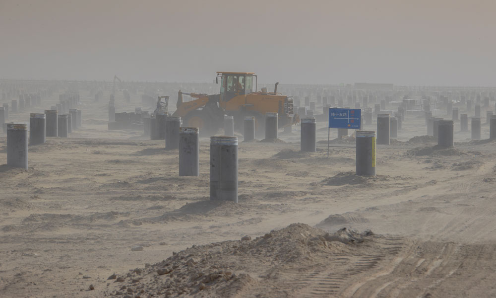 該項目作為塔里木油田公司目前施工的最大光伏發電項目，占地面積約2266公頃，南北、東西長寬均超過5公里。建設施工面廣線長，目前，已投入3500人到光伏面板架設、升壓匯集站及電化學儲能系統等建設工作，確保工程建設有序有效推進。圖為工程車輛在風沙中平整施工場地。 