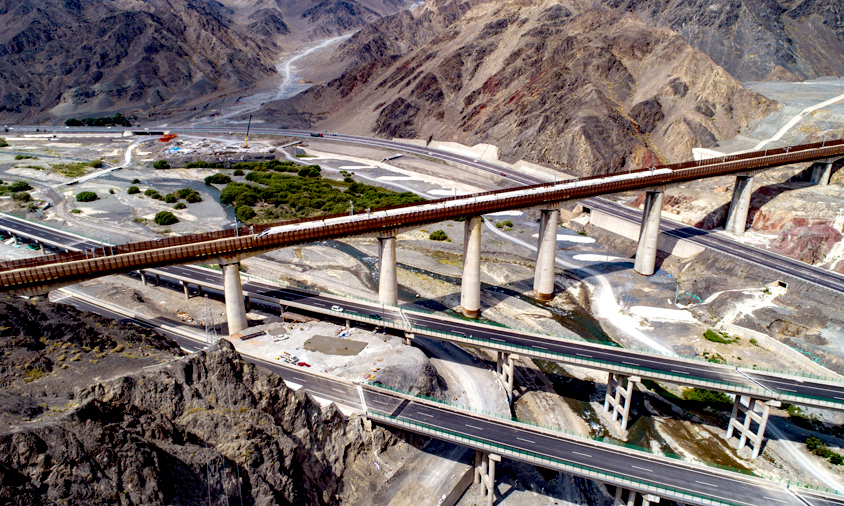 連霍高速公路起于連云港，止于霍爾果斯，全長4395千米，是“一帶一路”重要交通大動脈，中國最長的高速公路。小烏段橫跨亞洲最大的風(fēng)力發(fā)電站，穿越天山山脈，建設(shè)者克服常年大風(fēng)、低溫天氣及復(fù)雜的地質(zhì)條件，如期完成上行線通車任務(wù)。圖為連霍高速下穿蘭新高鐵。