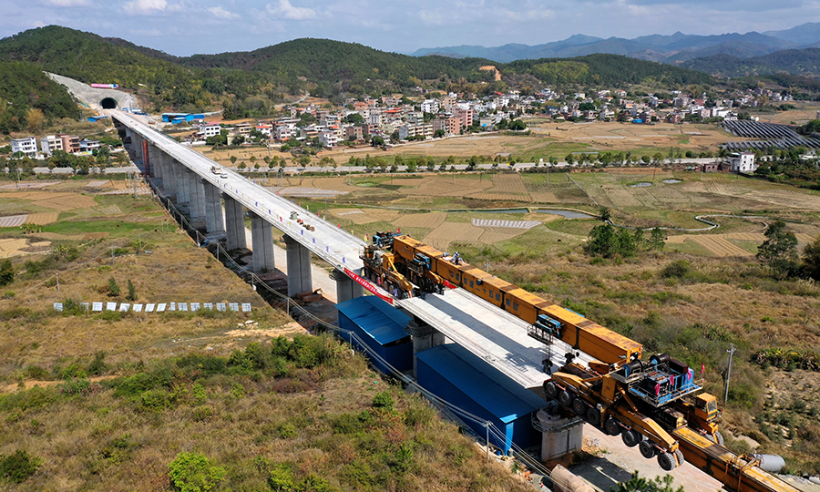 近日，國產(chǎn)WE-SC900型運架一體式架橋機前方伸縮支架不斷延伸，一榀820噸重、32米長的預(yù)制箱梁慢慢從尾部移動到“腹中”，穩(wěn)穩(wěn)落在贛深鐵路佳派跨G205國道特大橋的橋墩之間。粵港澳大灣區(qū)重點項目贛深鐵路項目架梁施工有序推進。圖為架梁施工俯瞰。（陳浩 攝）